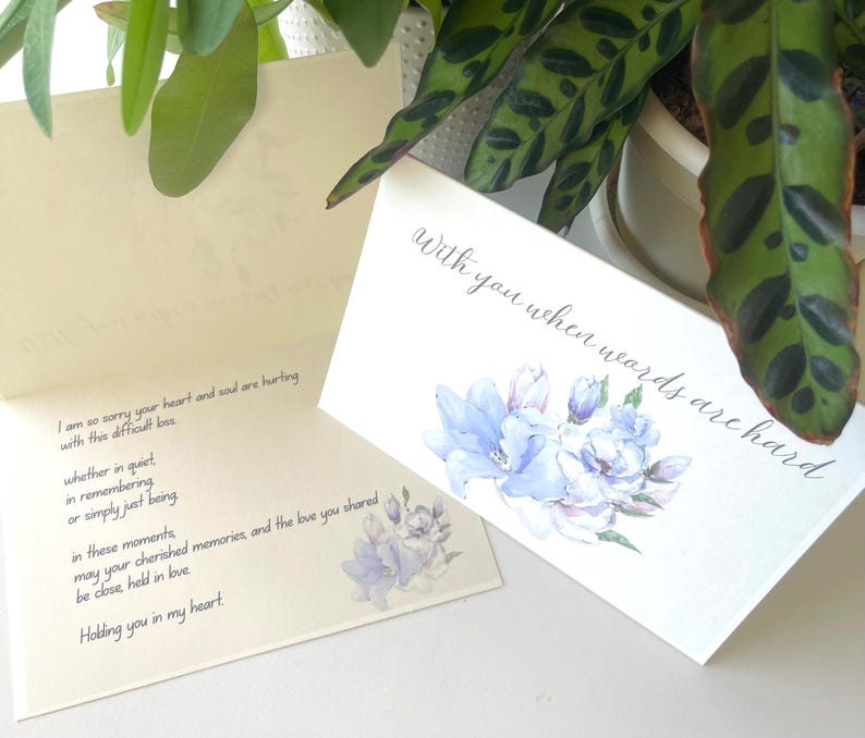 May include: A folded sympathy card with a white exterior featuring a watercolour illustration of blue flowers and the text "With you when words are hard." The interior is cream-coloured with black printed text expressing condolences and support.
