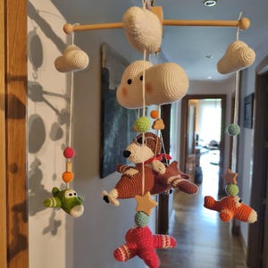 May include: A hanging mobile featuring a wooden cross frame with white crocheted clouds and several small crocheted airplanes in green, brown, orange, and pink. The airplanes are accented with small wooden stars and colorful crocheted spheres suspended by white strings.
