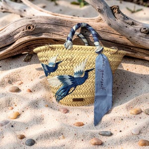 May include: Woven straw tote bag with blue and white embroidered bird design. The bag has a blue and white patterned handle and a blue ribbon with the text "love yourself". The bag is sitting on sand.