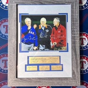 May include: Framed photograph featuring three men in jackets, with signatures below. The image is labeled "Legendary Texans" and includes the Texas Rangers logo. The frame is a light gray color.