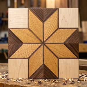 Könnte beinhalten: Ein quadratisches Wandkunstobjekt aus Holz mit einem geometrischen Sternmuster. Das Design besteht aus Segmenten aus hellem Ahornholz, mittelfarbener Eiche und dunklem Walnussholz, die in einem symmetrischen Sternmotiv angeordnet sind.