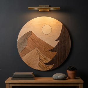 May include: A circular wooden wall art piece featuring a mountain landscape with a sun, crafted from various wood grains in light, medium, and dark brown tones. The artwork is illuminated by a brass-toned horizontal light fixture mounted above it.