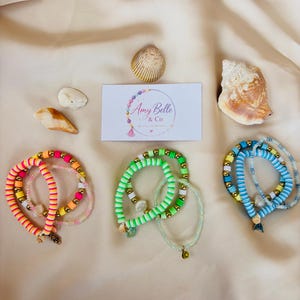 May include: Three sets of beaded bracelets featuring colorful clay discs, metallic gold beads, and small charms. The bracelets are arranged in groups of pink and orange, green, and blue tones, accompanied by three seashells and a business card.