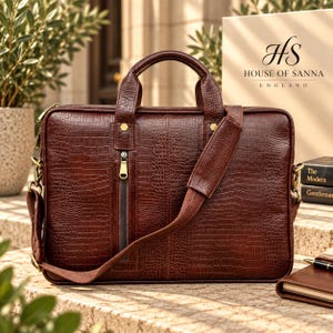 May include: A dark brown briefcase with a textured crocodile-embossed pattern, featuring two top handles, a detachable shoulder strap, and a vertical zippered front pocket with gold-tone hardware. The bag is branded with the House of Sanna England logo.