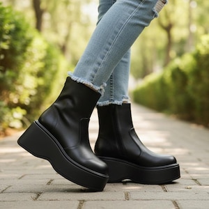 May include: Black platform ankle boots with a rounded toe and a chunky, angled sole. The boots have a side zip and are worn with light blue denim jeans. The boots appear to be made of leather.