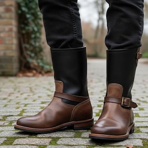 May include: A pair of brown leather boots with black uppers. The boots have a buckle closure and a strap around the ankle. The boots are worn with black jeans. The boots have a classic, rugged design.
