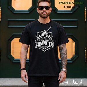 Peut inclure: T-shirt noir avec un motif blanc. Le motif indique "I AM THE COMPUTER" et représente une main tenant un tournevis, un éclair et un circuit imprimé. Le t-shirt est à col rond.