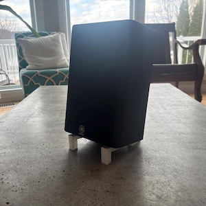 May include: A black Yamaha speaker on white stands, placed on a gray concrete table. The speaker is rectangular and has the Yamaha logo on the bottom. The background includes a window, a chair, and a couch.