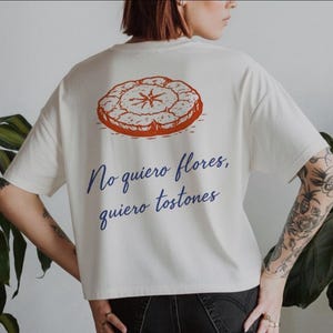 Könnte beinhalten: Cremefarbenes T-Shirt mit einer Grafik eines runden, orangefarbenen Lebensmittels auf der Rückseite. Unter der Grafik steht in dunkelblauer Schrift der Text "No quiero flores, quiero tostones".