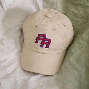 May include: A beige baseball cap featuring an embroidered logo on the front panel. The logo consists of the letters PR in red with a bold blue outline. The cap has a curved brim, a button on the crown, and stitched eyelets for ventilation.