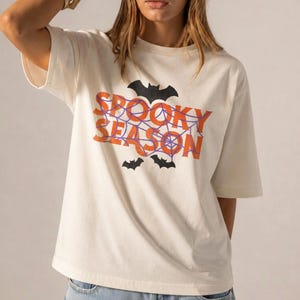 May include: An off-white short-sleeve t-shirt featuring the orange text SPOOKY SEASON layered over a purple spiderweb graphic. The design includes three black bat silhouettes positioned above and below the text, creating a graphic print.