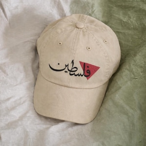 以下が含まれることがあります： ベージュの野球帽。黒い筆記体で「فلسطين」と赤い三角形が描かれています。柔らかい素材でできており、つばがカーブしています。クラシックスタイルです。
