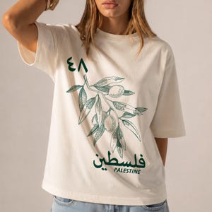 以下が含まれることがあります： オフホワイトの半袖Tシャツで、オリーブの枝のダークグリーンの線画が描かれています。デザインには左上にアラビア数字の48、枝の下にはアラビア文字と英語でPalestineという単語が含まれています。