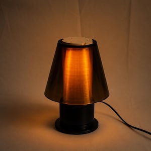 Könnte beinhalten: Eine moderne Tischlampe mit schwarzem Sockel und einem konischen, bronzefarbenen Schirm. Die Lampe ist beleuchtet und wirft einen warmen, orangefarbenen Schein. Ein schwarzes Netzkabel geht von der Basis aus. Die Lampe ist für den Innenbereich konzipiert.