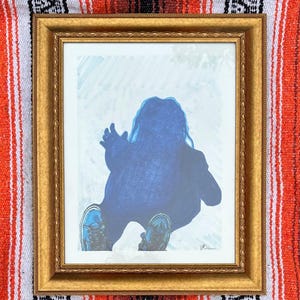 May include: A framed artwork featuring a blue silhouette of a person seated with arms raised. The figure is wearing shoes. The artwork is in a gold-colored frame, set against a white and blue background.