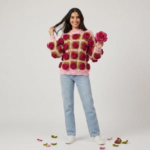 May include: A pink crochet sweater with burgundy rose appliqués. The sweater has a round neckline and long sleeves. The person is wearing light blue jeans and white sneakers. A large pink rose is held in the right hand.