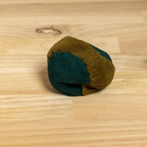 May include: A small, round footbag constructed from multiple panels of soft, textured material. The object features alternating sections of deep forest green and earthy olive brown, stitched together to form a compact, spherical shape.