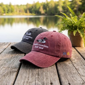 Könnte beinhalten: Zwei Baseballkappen mit gestickten Eistauchermotiven. Eine Kappe ist dunkelgrau, die andere weinrot. Die weinrote Kappe hat den Text "stupid canadian wolf bird" und die Zahlen "24 81" auf der Seite gestickt.