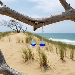 May include: Dangle earrings with a beach theme. Each earring features a stack of sea glass in shades of blue and white, suspended from a silver hook. The earrings hang from a weathered branch, with a sandy beach and ocean in the background.