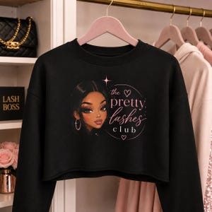 May include: Black cropped sweatshirt featuring a graphic of a woman's face and the text "the pretty lashes club" in pink. The design includes a star and hearts. The sweatshirt is on a pink hanger.