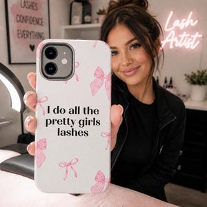 May include: White phone case with pink bow accents and the text "I do all the pretty girls lashes." The case is held up by a person. The background includes a neon sign that says "Lash Artist."