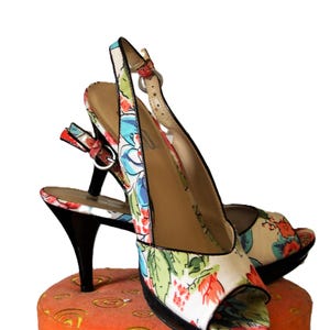 May include: A pair of floral print slingback heels with black heels and soles. The open-toe shoes feature a colorful pattern of red, blue, and green flowers on a cream background. The shoes are displayed on a multi-colored platform.