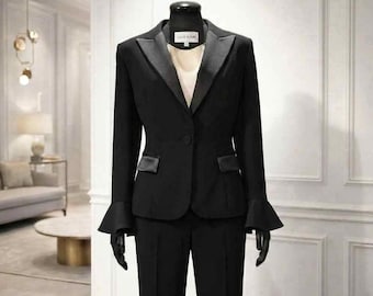 Puede incluir: Un conjunto de traje negro que incluye una americana con solapas de satén, cierre de un botón y puños acampanados. La americana se lleva sobre una prenda superior clara. El pantalón negro a juego tiene corte recto. La etiqueta dice LILIE RUSIN.