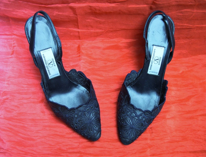Op de afbeelding: Een paar zwarte pumps met delicate bloemenborduursels. De schoenen hebben een spitse neus en een slingback-band. Het label op de schoenen luidt "Missy Valentino Garavani".