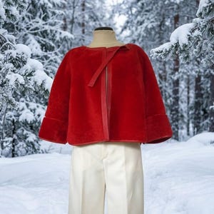 May include: A vibrant red cropped jacket with wide sleeves and a simple front closure, worn over a beige turtleneck sweater and cream-colored trousers. The jacket features a smooth, uniform texture and clean lines, providing a structured silhouette.