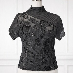 May include: A black short-sleeved top with a high mock neckline and a textured, grid-like fabric. The front panel features an asymmetrical overlay adorned with dense, shimmering black sequins arranged in a subtle, abstract pattern across the chest.