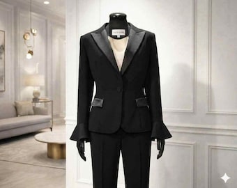 Op de afbeelding: Een zwart broekpak bestaande uit een blazer met satijnen revers, klepzakken en uitlopende mouwen, gecombineerd met een bijpassende broek. De blazer heeft een enkele knoopsluiting en wordt gedragen over een lichtbeige top. Het label binnenin toont LILLIE SUSAN.