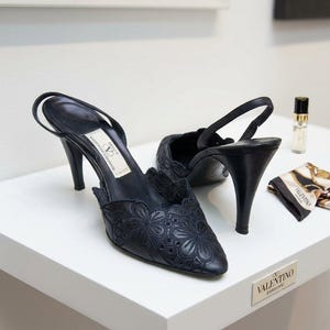 May include: A pair of black slingback heels featuring intricate floral embroidery and cut-out details on the pointed toe. The shoes have a high stiletto heel and a branded insole with the text Valentino Garavani. A small perfume bottle and a patterned silk scarf are nearby.