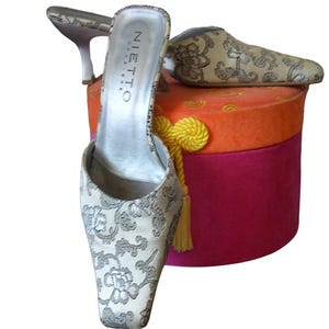 May include: A pair of cream-colored, open-toe high-heeled mules with a floral pattern. The shoes are displayed on a multi-colored, round box with a gold tassel. The brand name "NIETTO" is visible on the inner sole.