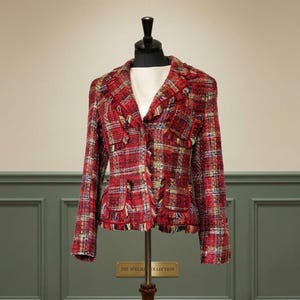 May include: A red plaid tweed blazer featuring a notched lapel, long sleeves, and two front pockets with flap closures. The garment is detailed with frayed edges along the lapels, pocket flaps, and hemline, showcasing a textured woven fabric.