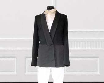 May include: A black blazer with a contrasting dark gray lower section, displayed on a mannequin. The blazer has a notched lapel, two buttons, and long sleeves. A white shirt and a cream-colored scarf are visible beneath the blazer.
