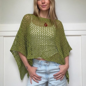 May include: An olive green crochet poncho featuring an open, loose-knit mesh pattern with a wide, draped silhouette. The garment has a boat neckline and short, wide sleeves that fall to the elbows, layered over a white top and light blue jeans.