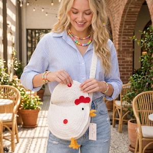 Puede incluir: Bolso bandolera blanco con forma de gallina de ganchillo, con detalles rojos y amarillos. El bolso tiene cierre de solapa y una correa larga. La persona lleva una camisa a rayas azules y blancas y vaqueros claros. También se ven joyas de cuentas de colores.