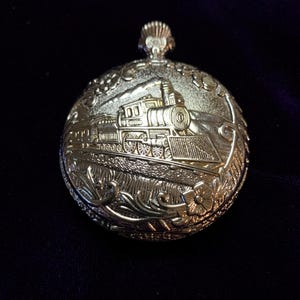 May include: A round gold-toned pocket watch case featuring an embossed design of a steam locomotive train on the front cover. The metal surface displays intricate floral detailing along the edges and a textured background behind the train engine.