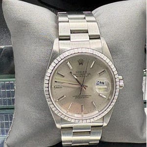 May include: A silver-toned Rolex Oyster Perpetual Datejust watch with a fluted bezel and a silver dial. The watch has a stainless steel bracelet and is displayed on a gray pillow. The watch face displays the date.
