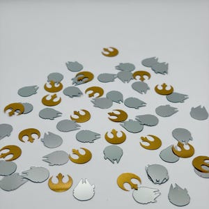 May include: Scattered gold and silver confetti in various shapes, including the Rebel Alliance symbol and the Millennium Falcon. The confetti is spread across a white surface, creating a festive and celebratory look.