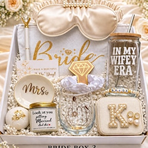 Op de afbeelding: Een bruidsgeschenkdoos gevuld met items met een bruiloftthema. Inclusief een slaapmasker, pen, bord met "Mrs" tekst, kaars, glas, sieradendoos met oorbellen en een beker. De doos is wit met gouden accenten.
