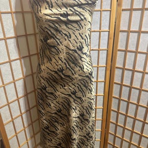 May include: A sleeveless, midi-length dress with a tiger stripe pattern in shades of tan and black. The dress features thin spaghetti straps and a cowl neckline. The fabric has a silky sheen.