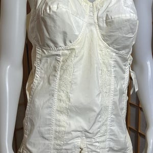 May include: A cream-coloured one-piece foundation garment featuring structured cups, vertical lace panelling, adjustable shoulder straps, and a side zip closure. The garment includes integrated garter clips at the bottom hem for hosiery attachment.