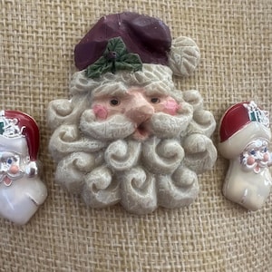 May include: A large Santa Claus face brooch with a dark red hat and green holly leaves, flanked by two smaller Santa face earrings featuring red hats, white beards, and silver-toned accents. The set displays detailed facial features and textures.