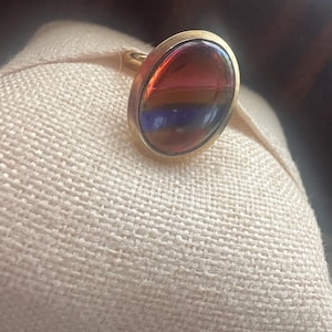 May include: A gold ring featuring a large, oval-shaped cabochon stone. The stone displays a smooth, polished surface with horizontal bands of deep red, orange, and dark blue colours, set within a simple, polished gold metal bezel setting.