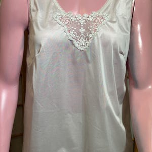 May include: A light beige camisole with a V-neckline and delicate white lace detailing. The camisole has a satin-like sheen and a simple, elegant design. The lace is floral-patterned.