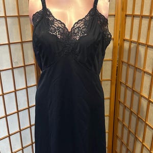 Op de afbeelding: Een zwarte slipdress met verstelbare bandjes en een kanten halslijn. De jurk wordt op een mannequin gepresenteerd en heeft een eenvoudig, elegant ontwerp. De jurk is gemaakt van een glad, zijdeachtig materiaal.