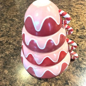 Op de afbeelding: Een gestapelde keramische maatbekerset in roze en rode tinten, die op een dessert lijkt. Elke beker is versierd met een wit golvend ontwerp en zuurstokhandvatten. Ontworpen voor gebruik in de keuken.
