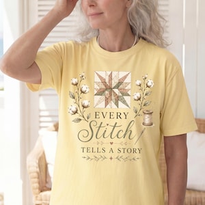 May include: A pale yellow short-sleeved t-shirt featuring a graphic print of a geometric quilt block, cotton sprigs, and a sewing spool. The text reads "EVERY Stitch TELLS A STORY" in a combination of serif and script fonts with decorative accents.
