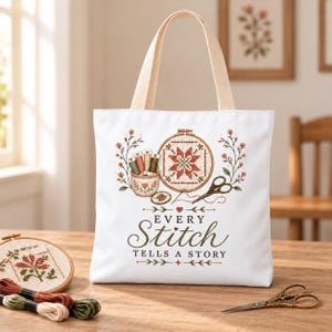 Könnte beinhalten: Eine weiße Tragetasche aus Segeltuch mit zwei beigen Henkeln und einem Aufdruck, der einen Stickrahmen, Stickgarn und eine Schere zeigt. Der Text "EVERY Stitch TELLS A STORY" steht in dunkelgrünen Buchstaben, umrahmt von floralen Akzenten.